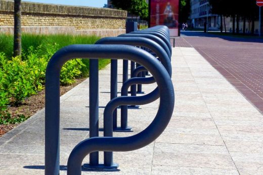 Supports vélos métalliques thermolaqués CRC alignés sur trottoir ensoleillé, proche zone végétale.
