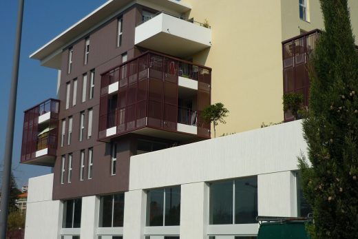 Bâtiment moderne, grandes baies vitrées, balcons rouges métallisés ; arbres et verdure devant.