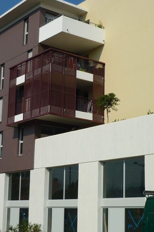 Bâtiment moderne, grandes baies vitrées, balcons rouges métallisés ; arbres et verdure devant.